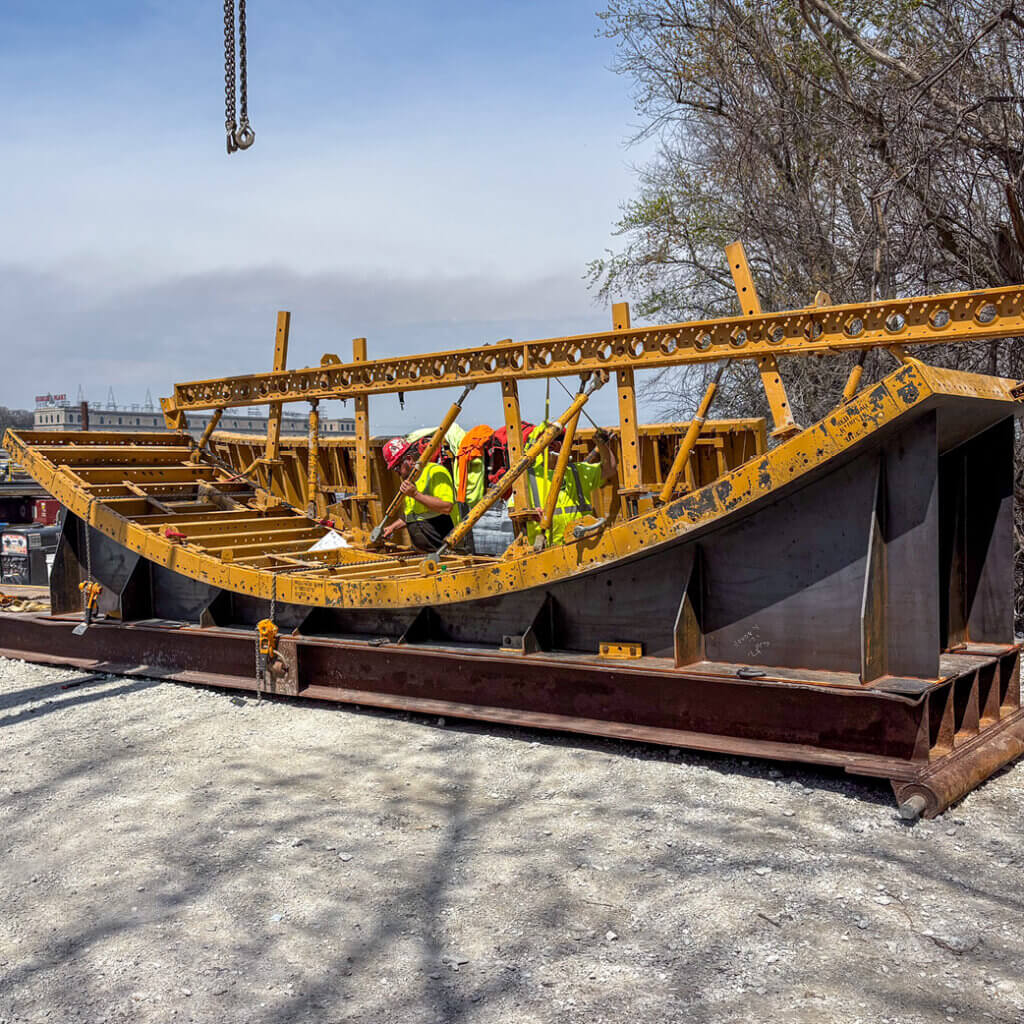 Crews finishing the steel face of a REDI-RADIUS® spillway form in the yard. Shaped just once, it holds the ogee’s radius pour after pour, with the E-BEAM & SUPER STUD® ‘skeleton’ keeping the assembly true from the yard to the barge.